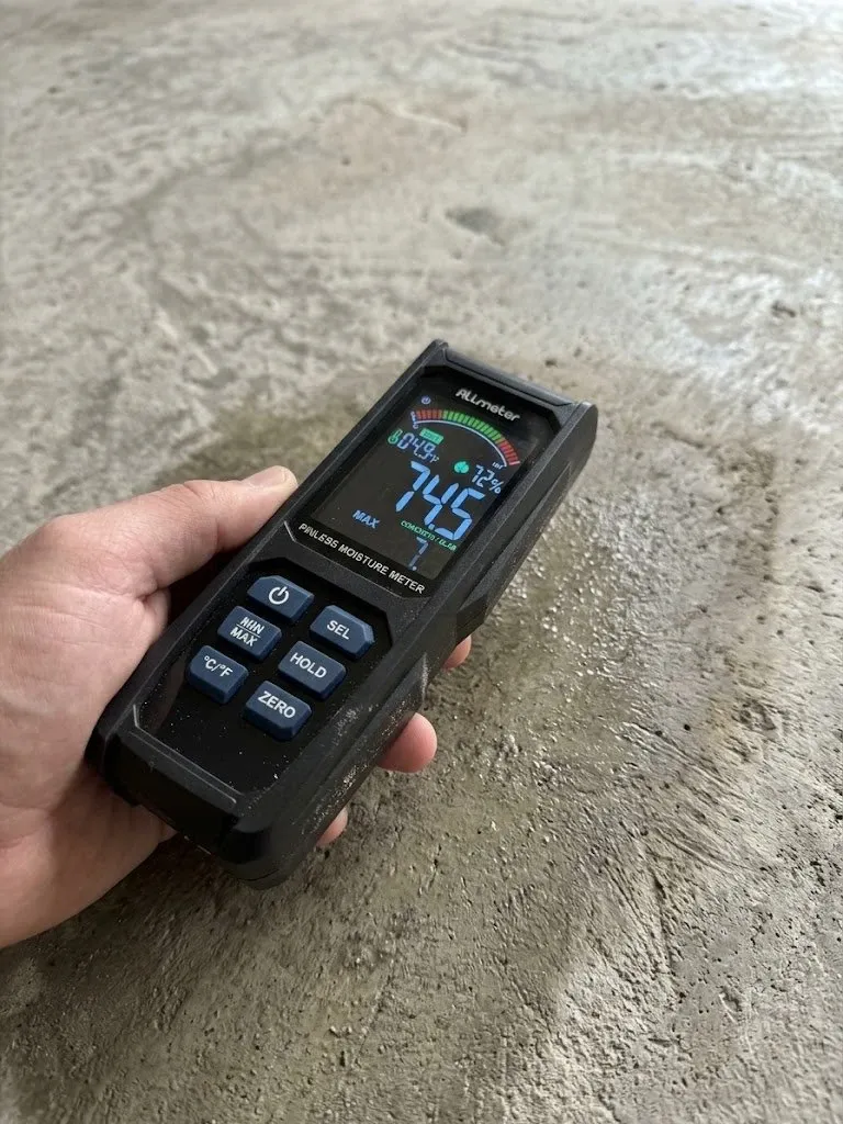 Hand holding a pinless moisture meter with its screen lit, at a job site