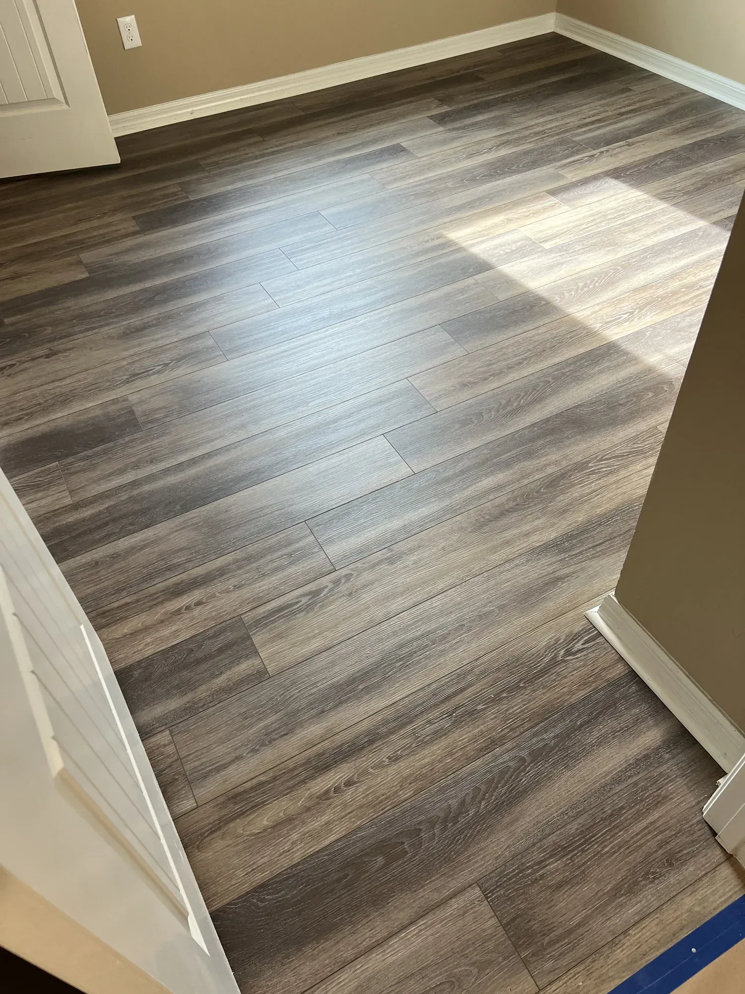 Corner of a room with newly installed grey wood-look plank flooring and a length of new baseboard resting against the wall