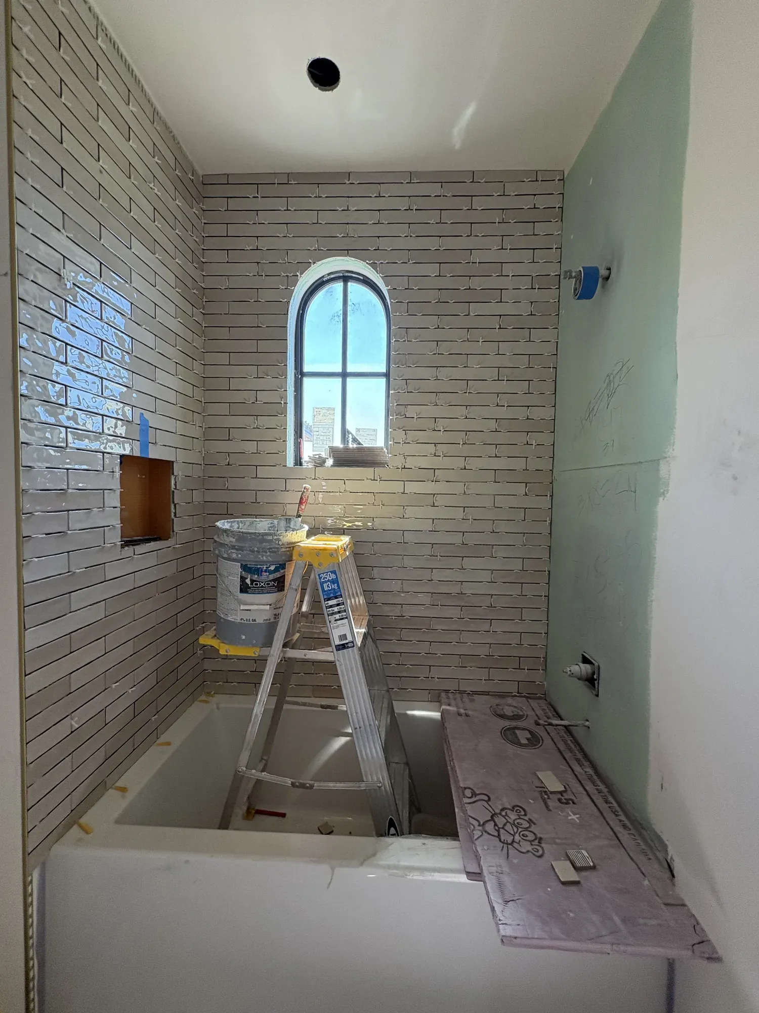 Bathroom mid-installation, with narrow wall tile going up around an arched window, spacers still in place and the tub protected