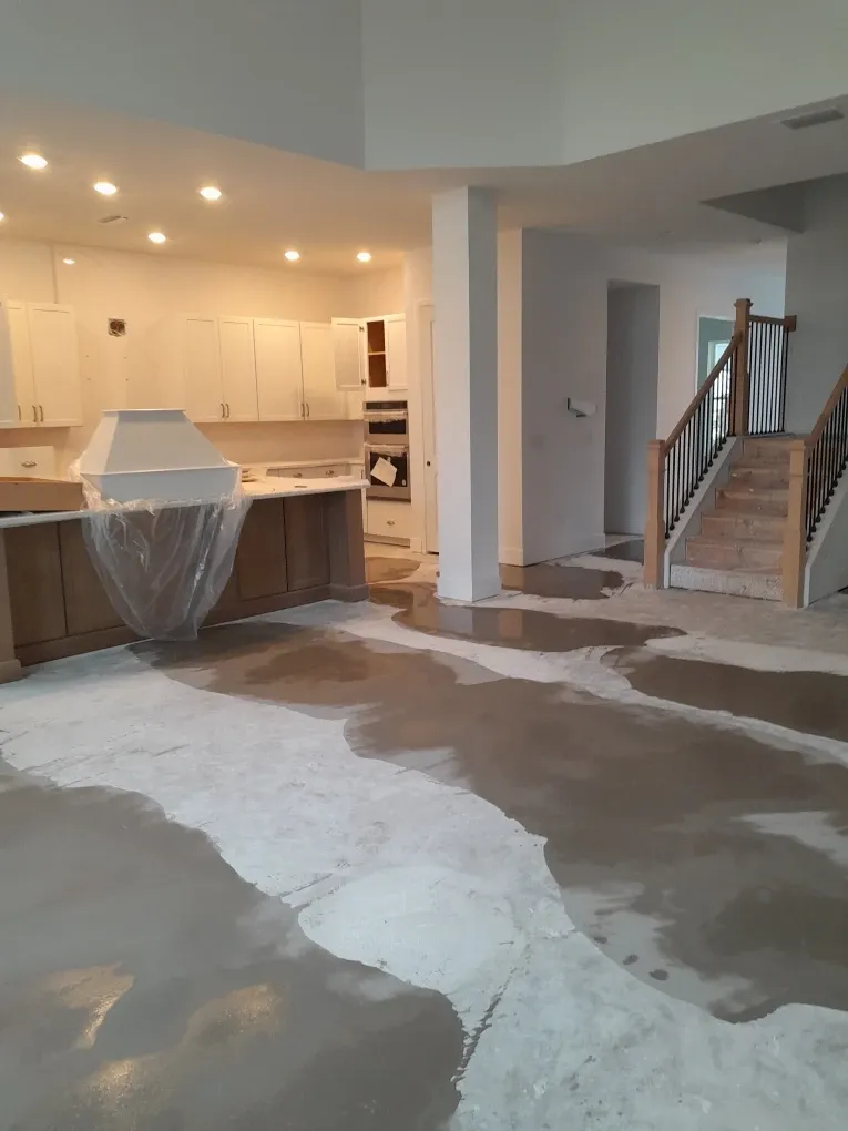 Open kitchen and living area with the slab patched in wide swathes of levelling compound, the range hood still wrapped in plastic and a finished staircase to one side