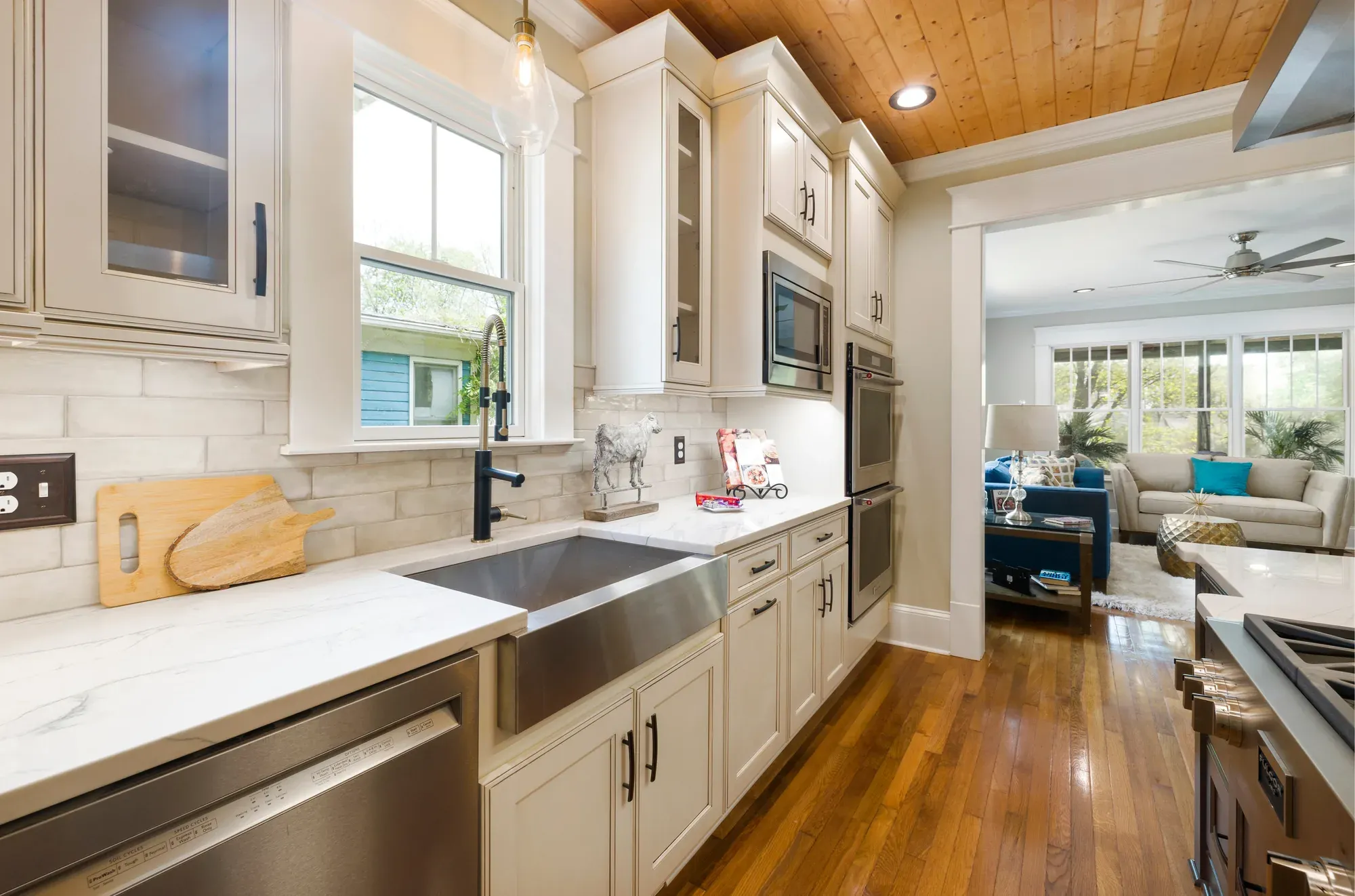 A cream shaker kitchen with a farmhouse sink, glass-front cabinets and a light countertop, opening onto a living room