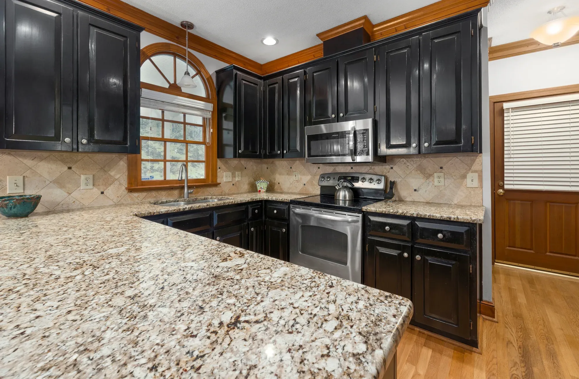 A kitchen with dark wood cabinets, stainless steel appliances and a warm beige granite countertop wrapping around the corner counter