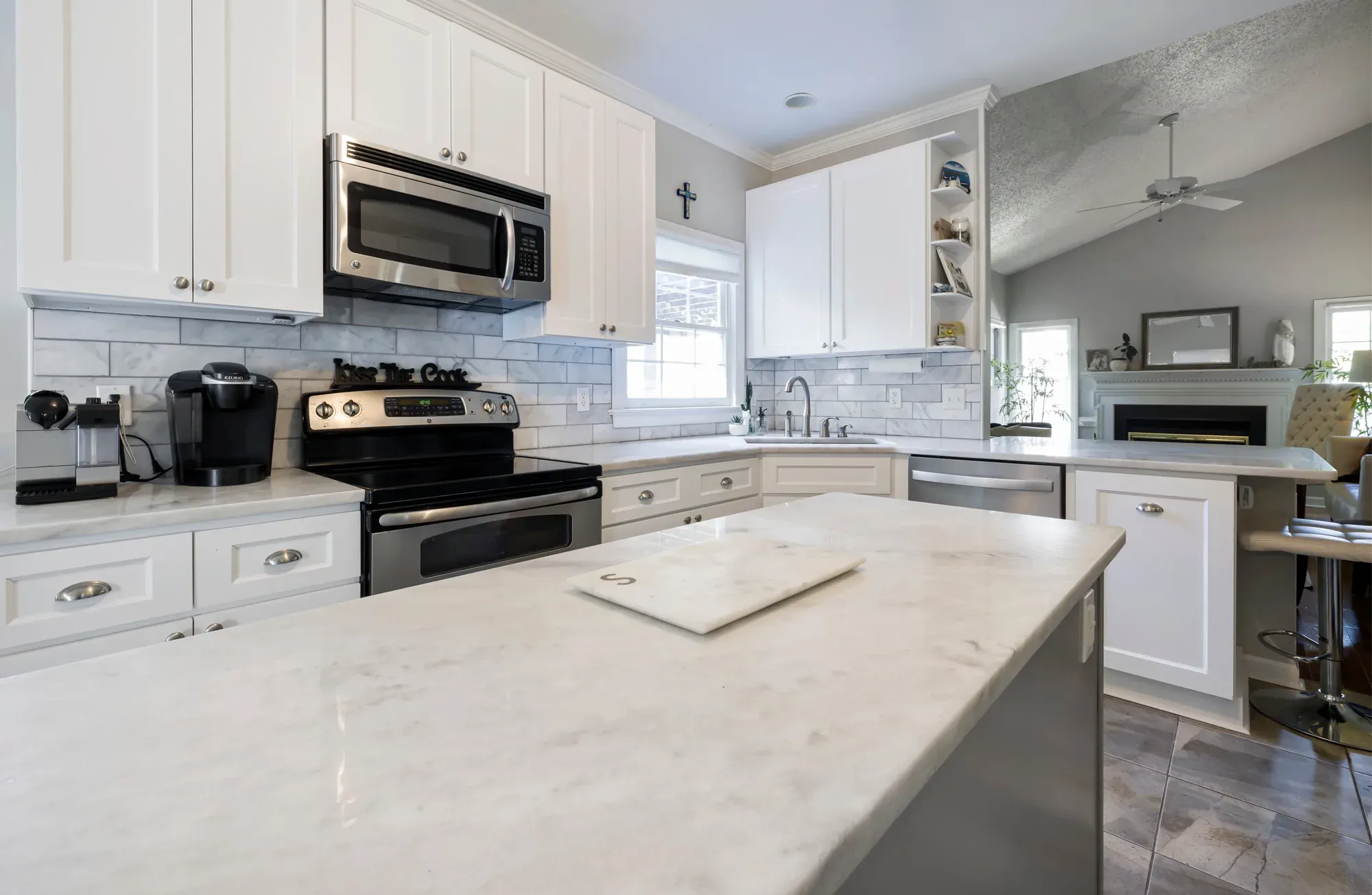 A white kitchen with an island topped in light veined stone, a cutting board on the surface and a stainless steel range against the back wall