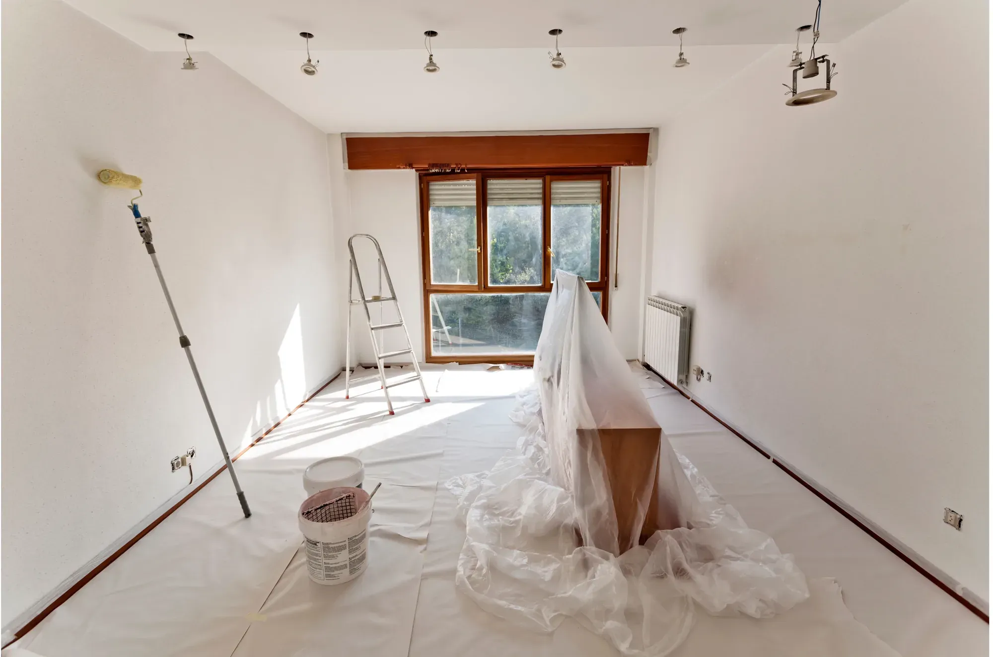 An empty room prepared for painting, with drop cloths over the floor, furniture covered in plastic and a roller on an extension pole leaning against the wall