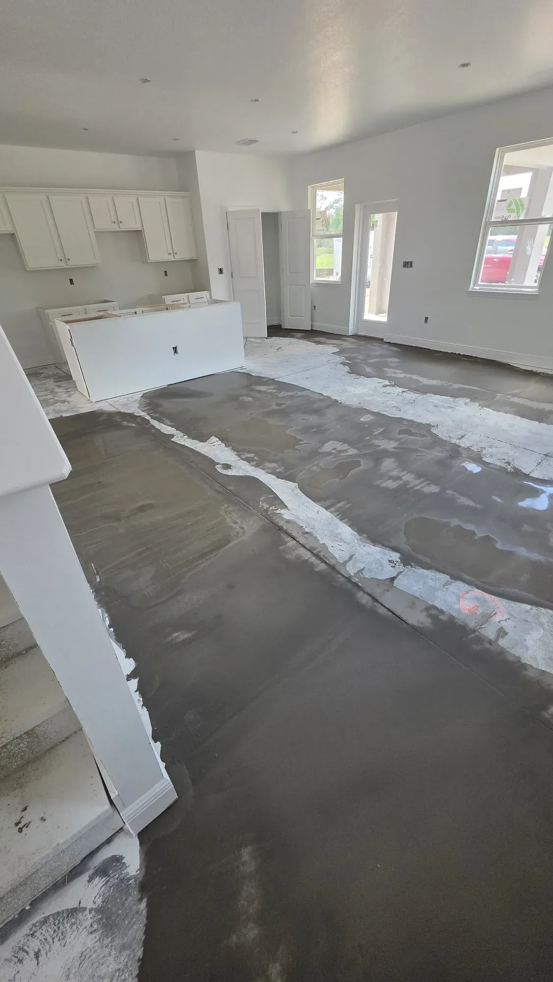 Open-plan room in a house under construction, with self-leveling compound spread over the slab and drying in patches, white kitchen cabinets already hung along the far wall