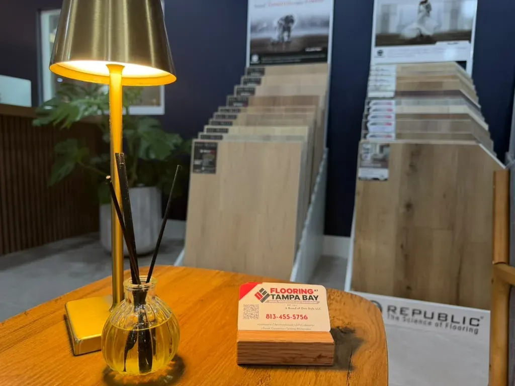 A wooden table in the showroom with a small gold lamp, a glass vase and a stack of Flooring at Tampa Bay business cards, with flooring sample boards on stands behind