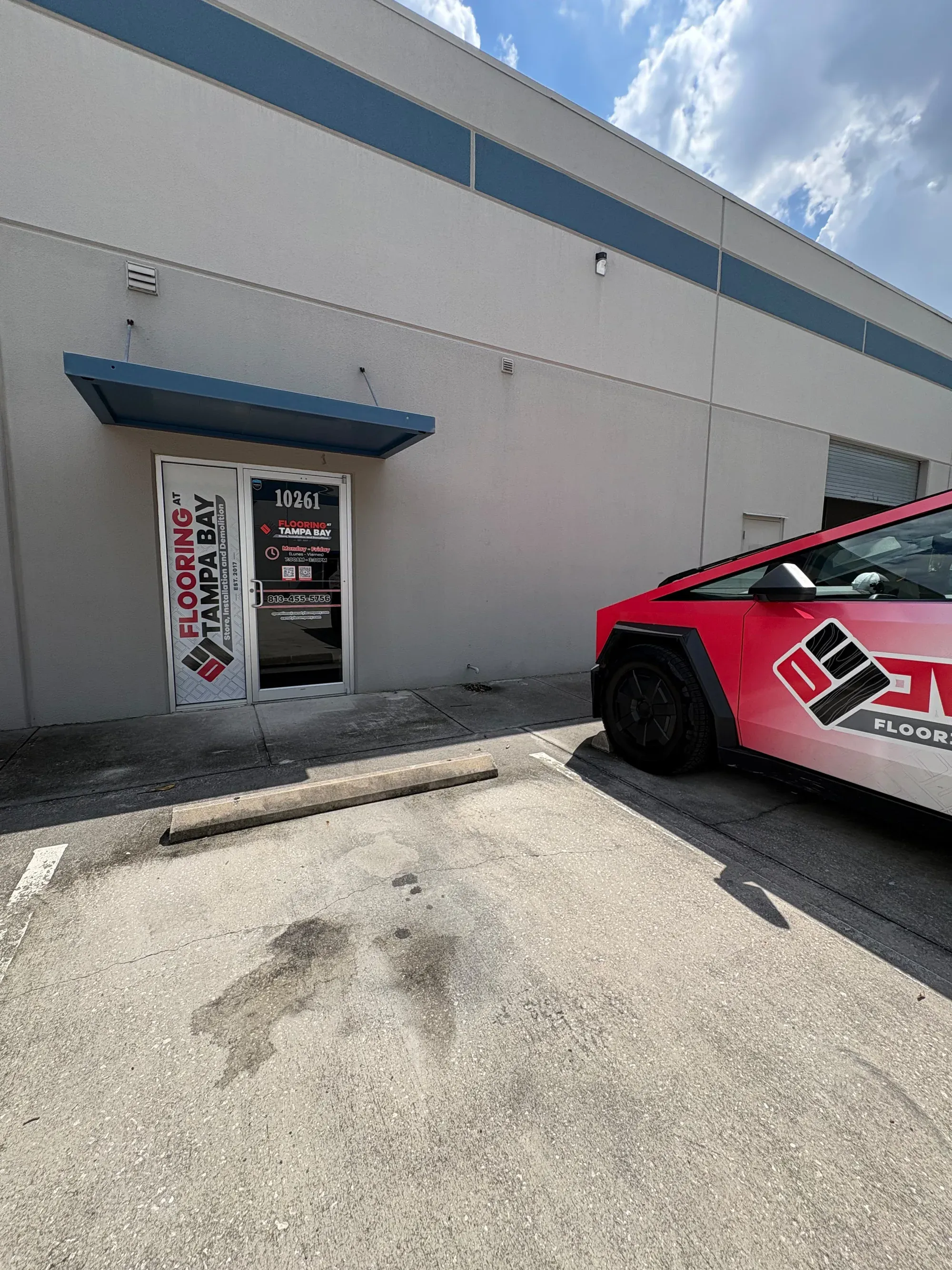 The outside of the showroom: the entrance door with the Flooring at Tampa Bay sign and the street number 10261 over it, and the paved parking lot in front with a company-wrapped car