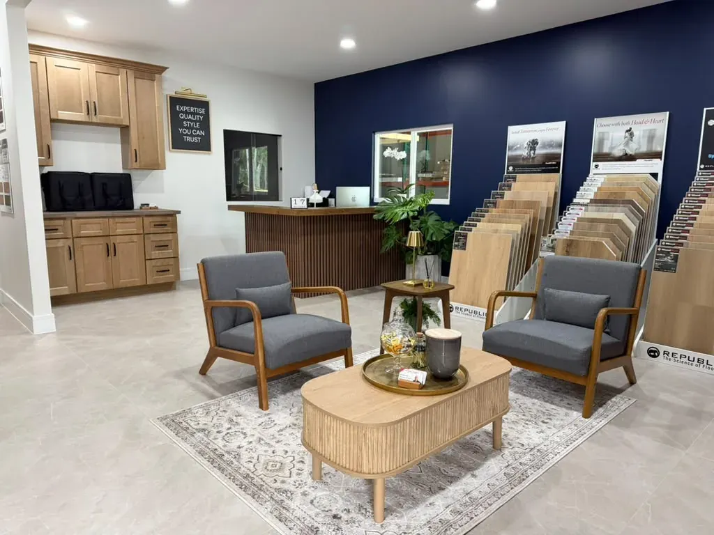 The showroom counter in light wood with a glass display case behind it, two gray armchairs and a round wooden table with a tray, and sample stands to the right
